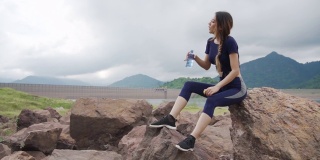 疲惫的年轻美丽的亚洲女运动员穿着运动服坐在岩石上，从一个瓶子里喝水后在山道跑步。运动女孩跑步者放松和享受美丽的自然。户外运动健身概念。