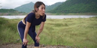 疲惫的年轻亚洲妇女在草地上跑步之间深呼吸的慢动作。汗流浃背的运动女性在草地上慢跑进行有氧训练。户外运动的生活方式和极限运动训练的概念。