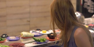在一家亚洲咖啡馆里，一名年轻女子用火锅做饭。食物被盛在传送带上的一个小盘子里。食品输送机。