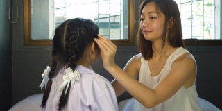 亚洲母亲在她的卧室里穿着校服给女儿做头发，准备早上上学。