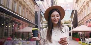 迷人的肖像迷人的快乐的年轻女子戴着时髦的帽子，走在现代建筑与街头咖啡馆之间，一只手拿着咖啡和电话在另一个