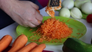 一个男人用研磨器磨胡萝卜。附近的蔬菜，烹饪南瓜鱼子酱。桌上放着西葫芦，胡萝卜，洋葱和西红柿。特写镜头。视频素材模板下载