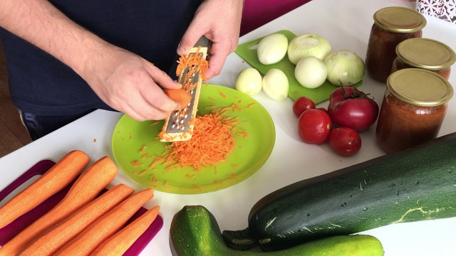一个男人用研磨器磨胡萝卜。附近的蔬菜，烹饪南瓜鱼子酱。桌上放着西葫芦，胡萝卜，洋葱和西红柿。