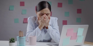 一名亚洲女性在使用笔记本电脑工作时发烧，打喷嚏，坐在家里的办公室里。