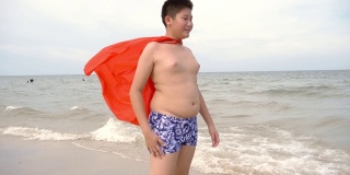 快乐的亚洲男孩披着红布披肩站在海滩上，慢镜头。