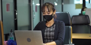 亚洲女员工戴口罩，在商务办公室使用笔记本电脑，与同事保持社交距离，以保护冠状病毒或covid-19疫情大流行。职场新常态政策