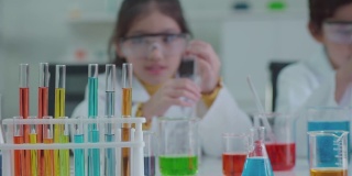 穿着科学家制服的亚洲孩子们玩得很开心。学习科学实验室实验试管玻璃水在桌子上的各种颜色，聚焦试管颜色背景模糊