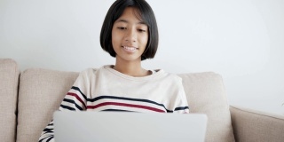 手持。快乐的亚洲小女孩在笔记本电脑上视频通话，并在上午坐在家里客厅的沙发上与导师招手打招呼