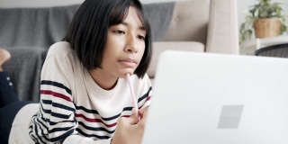 手持。亚洲女孩在网上学习通过网络在笔记本电脑上和导师，躺在客厅的地板上。在家在线学习的概念