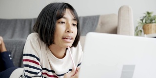 亚洲女孩在网上学习通过网络在笔记本电脑上和导师，躺在客厅的地板上。在家在线学习的概念