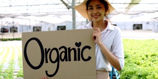 近距离观察年轻的亚洲女士农民看相机拿海报纸板与字有机靠近柜台与新鲜蔬菜。健康饮食的概念