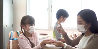 年轻女教师在教室里用酒精喷雾器消毒学生的手。亚洲妇女戴口罩，用洗手液清洁学生的手。学校在隔离和封锁后重新开学