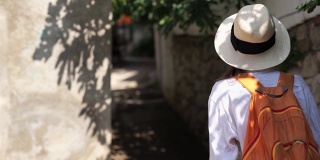 镜头跟随美丽性感的女孩穿着牛仔裤短裤，草帽和白衬衫上楼在狭窄的街道