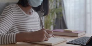 亚洲女孩的手正在做作业和使用平板电脑。亚洲妇女戴着防护口罩在家感染流感或COVID-19。健康与疾病概念