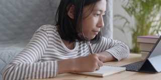 手持。一个亚洲女孩正在做作业，早上在桌子上用平板电脑。在家在线学习概念