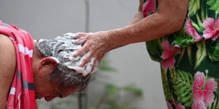 亚洲华人老年妇女洗她的丈夫的头发，洗头在前院当没有水供应