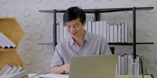 一名亚洲高级男子正坐在一间办公室里，看着一台笔记本电脑，面带微笑地工作着，笑容满面，事业成功，在家工作