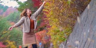 慢动作的微笑年轻美丽的亚洲女子游客扔红色枫叶在秋天的公园。快乐漂亮的女孩，放松和享受美丽的自然。日本旅游度假和秋季季节改变概念。