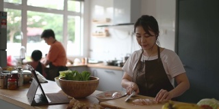 亚洲华人母亲在厨房柜台准备食物，并使用数字平板电脑通过互联网学习