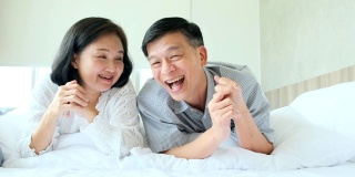 幸福安康健康的老亚洲夫妇笑着一起笑着享受躺下休闲在床上，快乐的老夫妇躺在床上看垫相机慢镜头