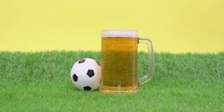一杯清凉的泡沫啤酒站在草坪上，黄色的背景。小玩具足球滚了出来。人拿起杯子解渴，又放回去