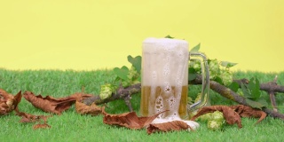 大杯子里装满了啤酒，厚厚的泡沫，黄色的底色。杯子放在绿色的人造草坪上，上面装饰着干树叶、树枝和一簇簇啤酒花