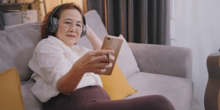 亚洲资深女性坐在沙发上，戴着无线耳机听在线音乐，而在家放松。在客厅听音乐或播客。