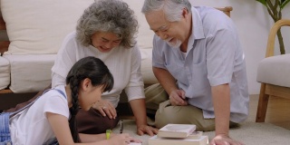 亚洲家族的祖父坐在客厅里笑着，用手摸着头表示爱。看孙女躺着写书，幸福老年退休