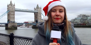 英国伦敦塔桥附近，年轻的女记者戴着圣诞帽，用麦克风讲话