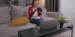 广角拍摄和手持:迷人的亚洲高级女性通过笔记本电脑学习在线摄影课。坐在家里客厅的沙发上和她的老师学习如何用视频通话技术使用数码相机。