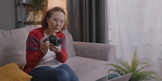 广角拍摄和手持:迷人的亚洲高级女性通过笔记本电脑学习在线摄影课。坐在家里客厅的沙发上和她的老师学习如何用视频通话技术使用数码相机。