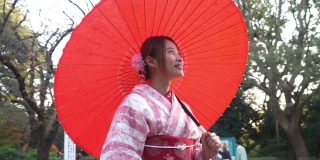 微笑的年轻美丽的亚洲女性游客穿着粉红色的传统日本和服拿着红色的伞放松和享受旅游假期在东京城市的公共公园，日本