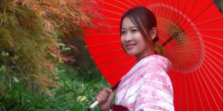 微笑的年轻美丽的亚洲女性游客穿着粉红色的传统日本和服拿着红色的伞放松和享受旅游假期在东京城市的公共公园，日本