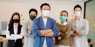 由于Covid-19流感大流行和防护，小型企业创业概念，亚洲商界领袖带着口罩微笑，与商业团队交叉臂自信地站在共同办公空间