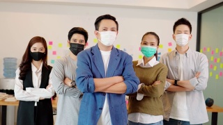 由于Covid-19流感大流行和防护，小型企业创业概念，亚洲商界领袖带着口罩微笑，与商业团队交叉臂自信地站在共同办公空间视频素材模板下载