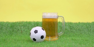 将冰凉爽口的泡沫啤酒倒进大杯子里，杯子立在人造草坪的绿草上，黄色背景，正面观景。小玩具足球滚了出来。