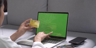 这位年轻的女士正在用银行卡进行网络支付，坐在家里的椅子上用笔记本电脑工作。网上购物，互联网和现代科技的概念。
