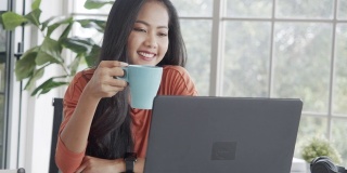 亚洲女商人微笑着喝咖啡，用笔记本电脑与她的团队在隔离期间进行视频通话。
