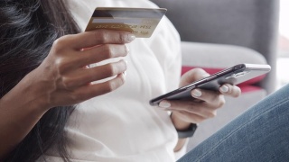 手握信用卡和智能手机的年轻亚洲女性顾客坐在家里的沙发上。视频素材模板下载