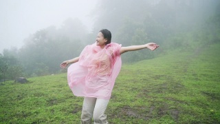 快乐的年轻亚洲女人旅行者在粉红色的雨衣享受和有乐趣的旅行美丽的山在雨中。微笑着的漂亮女孩感到自由，在雨中跳舞。视频素材模板下载