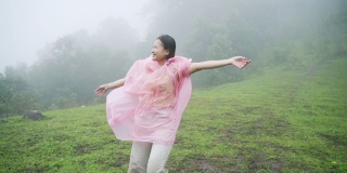 快乐的年轻亚洲女人旅行者在粉红色的雨衣享受和有乐趣的旅行美丽的山在雨中。微笑着的漂亮女孩感到自由，在雨中跳舞。