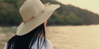 迷人的亚洲女人戴着软呢帽站在海滩上看海景美丽的日落。