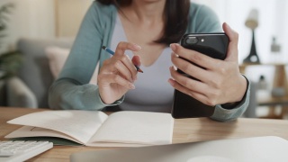 亚洲女性自由职业者手使用智能手机检查电子邮件，阅读商业报告，同时在笔记本上做笔记在家庭办公室工作视频素材模板下载