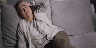 资深亚裔女性坐在家里的沙发上，对自己的生活感到严肃和担忧。老年女性生病或头痛。