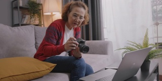 中景和手持式:迷人的亚洲高级女性通过笔记本电脑学习在线摄影课程。坐在家里客厅的沙发上和她的老师学习如何用视频通话技术使用数码相机。