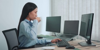 一名亚洲妇女在清晨喝咖啡，同时在思考电脑屏幕上的程序和代码。