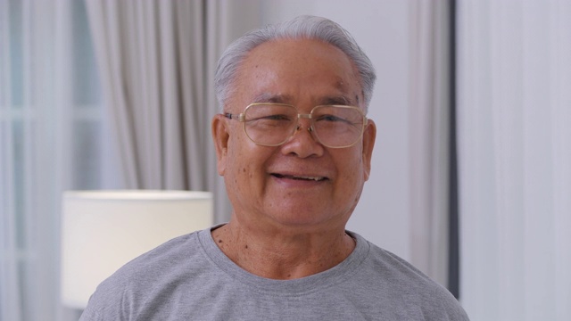 亚洲年长的白发老人在家里看着照相机。快乐善良的老人在一个好心情微笑。快乐的长者成熟的祖父带着牙齿的微笑在家里或退休的房子。高级的肖像