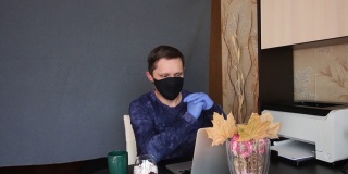 一名戴着面具和手套的男子通过视频链接进行交流。坐在桌子前的笔记本电脑前。他身后伸展着一块灰色的背景布。甜点和咖啡就在附近。