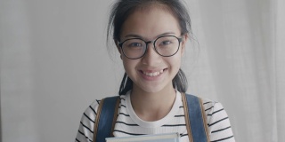 亚洲女人的肖像戴眼镜，快乐和微笑拿着书背包去学校，看着相机