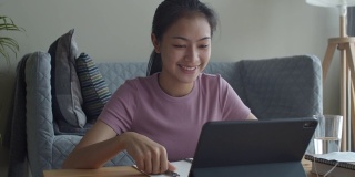 快乐的亚洲女性在家里的客厅里用平板电脑向在线家教挥手或打招呼。提出在家在线学习的概念